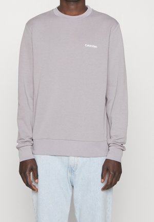 Persona che indossa una felpa Calvin Klein di un viola chiaro e jeans blu chiaro, in piedi contro uno sfondo bianco semplice.
