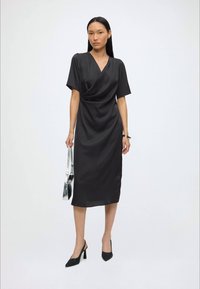 Žena v čiernych midi zavinovacích šatách, čiernych slingback lodičkách, drží striebornú kabelku, stojí na svetlom jednoduchom pozadí.