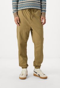 Pantaloni kaki cu talie elastică, buzunare laterale, margini manșonate și o croială lejeră. Asortați cu adidași albi cu accente negre.