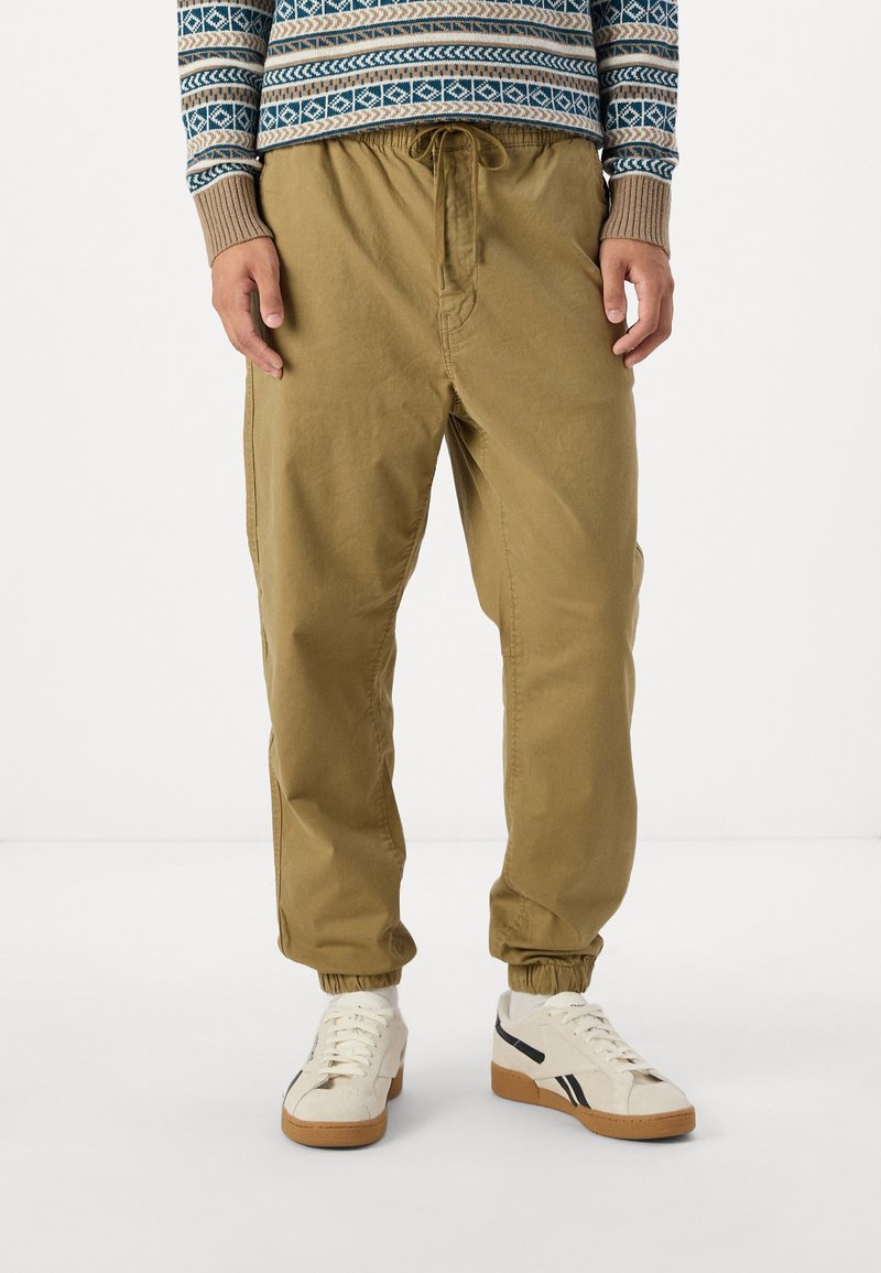 Pantaloni kaki cu talie elastică, buzunare laterale, margini manșonate și o croială lejeră. Asortați cu adidași albi cu accente negre.