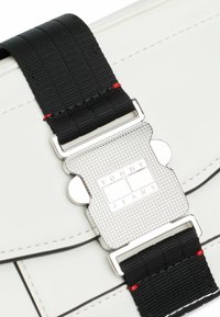 Bolso blanco con una correa negra texturizada y una hebilla plateada, que presenta el logo "TOMMY JEANS" en un diseño rectangular.