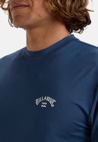Camisa corta de manga azul hecha de una tela suave, con un logo blanco de "Billabong" en el pecho y un escote redondo con detalle de costura.