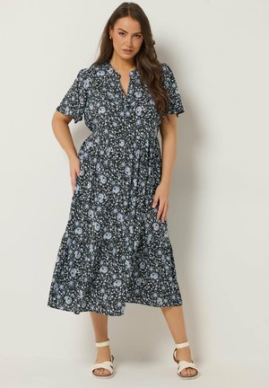 Kvinde iført en sort midi-kjole med hvidt blomsterprint og korte ærmer, kombineret med hvide ankelremssandaler, står op ad en neutral væg.