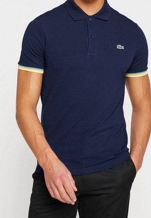 Man draagt een marineblauw poloshirt met geel-witte gestreepte mouwranden en een klein groen krokodillenlogo op de borst.