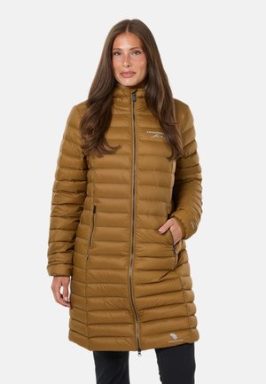 Femme portant une longue veste matelassée marron avec fermeture éclair à l'avant, debout avec une main dans la poche devant un fond uni.