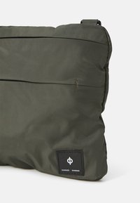 Borsa imbottita di colore verde oliva con una superficie liscia, dotata di una tasca con zip frontale e di una toppa nera con logo bianco.