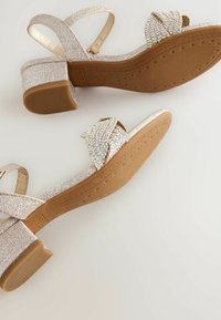 Tacones plateados brillantes con un bajo tacón bloque, un diseño de tiras cruzadas adornado con strass y una suela de goma texturizada para mayor agarre.