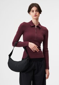 Schwarze Schultertasche aus weichem Stoff, gebogene Form, mit zwei runden Akzenten. Das Modell trägt einen burgunderfarbenen Reißverschluss-Oberteil und schwarze Hose.