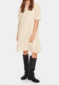 Kermakudonne, jossa on siluetteja, lyhyet hihat ja aaltoileva helma. Yhdistetty polveen saakka ulottuviin mustiin nahkasaappaisiin.