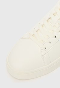 White leather sneaker with a smooth texture, rounded toe, and textured rubber sole; features flat laces and minimal stitching details.