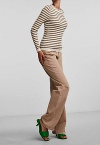 Osoba w beżowych spodniach, beżowo-białej, paskowanej bluzce z długim rękawem, i jaskrawych, zielonych sandałach na obcasie, stojąca z lekko ugiętą nogą.