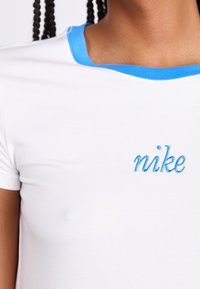 Bílé bavlněné tričko s krátkými rukávy, modrý žebrovaný límec a modré vyšívané logo "nike" na přední straně. Měkká textura, přiléhavý střih.