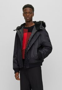 Chaqueta de plumas negra con capucha forrada de piel, que presenta un cierre de cremallera y botones, además de puños acanalados y una textura suave y brillante.