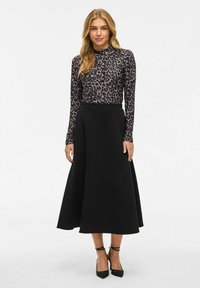 Femme aux cheveux longs blonds portant un haut à manches longues en imprimé léopard, une jupe midi noire et des talons à bride de cheville noirs, se tenant sur un fond blanc uni.