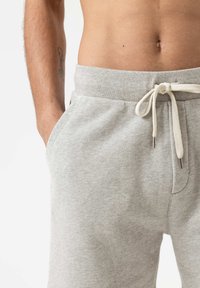 Pantalons de survêtement gris clair en tissu doux, avec une taille élastique à cordon, des poches latérales et une coupe décontractée. Texture lisse et design simple.