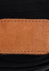 Rechthoekige beige leren patch met zichtbare stiksels op een zwarte gestructureerde stofachtergrond.