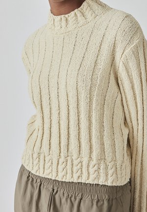 Kremowy sweter z dzianiny prążkowanej z teksturowanymi pionowymi paskami i warkoczowym wzorem u dołu, noszony z khaki spodniami.