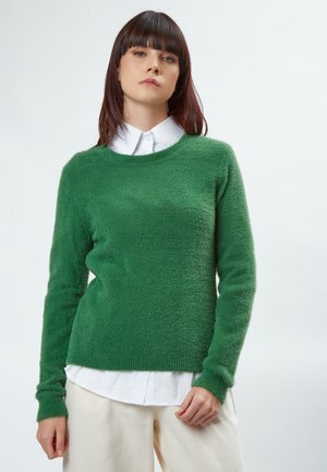 Maglione verde e morbido con maniche lunghe e scollatura rotonda, indossato sopra una camicia bianca con colletto, abbinato a pantaloni di colore chiaro.