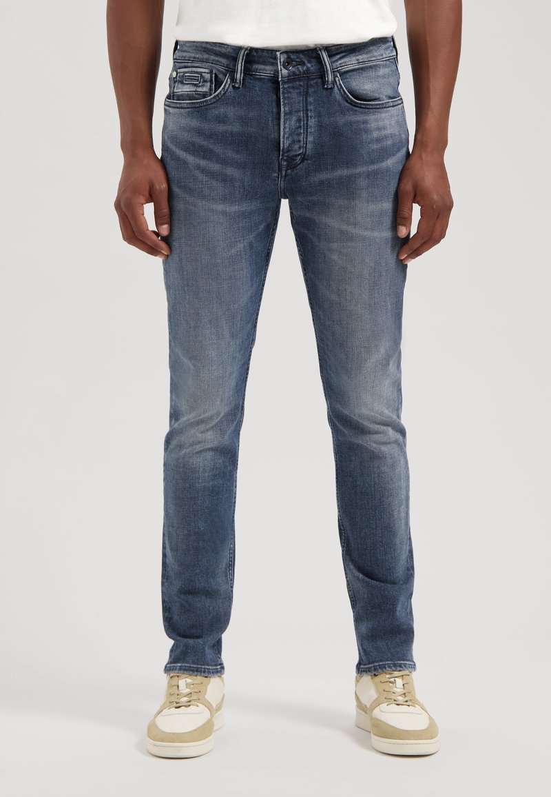 Homme portant un jean slim bleu délavé et des baskets beige-blanc, debout devant un fond clair uni.
