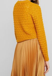 Žena v hořčicově žlutém pleteném svetru a skládané bronzové saténové sukni, zobrazená z boku a zezadu, na neutrálním pozadí.