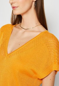 Pull col en V de couleur orange vif, texture tricotée, manches courtes avec motif en treillis sur les épaules, accompagné d'un collier métallique fin.