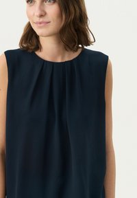 Marineblaues ärmelloses Oberteil mit Rundhalsausschnitt, das an der Vorderseite weiche Falten aufweist. Hergestellt aus leichtem Material mit einer glatten, matten Textur.