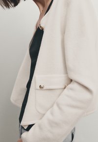 Cream-colored knit jacket with a cropped design, featuring a gold button, side pocket detail, and soft texture. Worn over a black top.