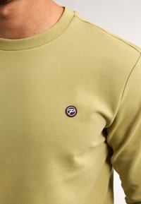 Sudadera verde oliva en una suave mezcla de algodón con un cuello redondo acanalado. Presenta un parche de logo redondo en negro y rojo en el pecho.