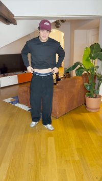 Sweatshirt oversized preto, calças largas pretas e ténis prateados. O fundo inclui um sofá castanho e uma planta verde, com pavimento de madeira.