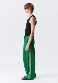 Groene wijde broek met gouden zigzagaccenten aan de zijkant, gecombineerd met een zwart tanktopje. Het model draagt een zonnebril en zwarte schoenen.