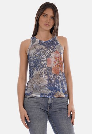 Donna con lunghi capelli castani che indossa un top blu senza maniche con grandi stampe floreali e jeans grigio chiaro, rivolta in avanti su uno sfondo semplice.