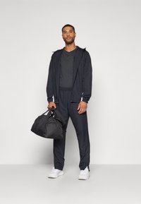 Tenue de sport pour homme en noir, comprenant une veste zippée, un pantalon assorti et des baskets blanches, tenant un sac de sport noir. Texture lisse.
