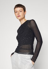Musta pitkähihaista puseroa, jossa on läpinäkyvää materiaalia ja ripoteltuja pieniä strasseja sekä mukautuva muoto, on yhdistetty hopeisiin pantsuihin.