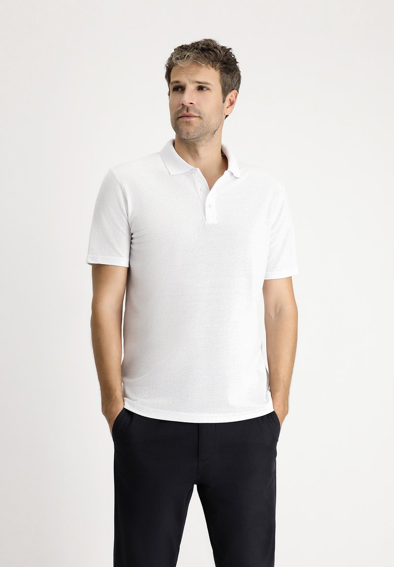 Biała koszulka polo z teksturowanej tkaniny, z krótkim rękawem i zapięciem na trzy guziki, w połączeniu z ciemnymi spodniami. Minimalistyczny design.