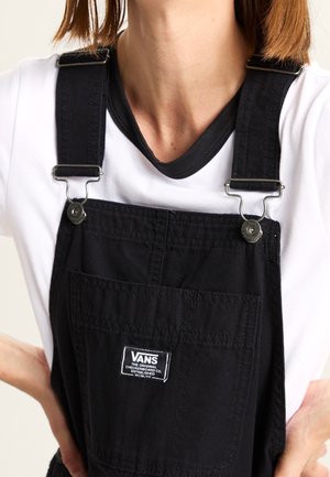 Persoon draagt zwarte Vans-overalls met zilveren gespen over een wit T-shirt, handen rusten op de heupen, bijgesneden ter hoogte van nek en taille.
