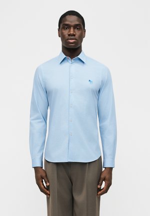 Muž v svetlomodrej košeli s dlhým rukávom a malým vyšívaným logom, v kombinácii so sivými nohavicami, stojaci na jednofarebnom pozadí.