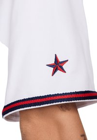 Camicia di cotone bianca con orlo rotondo. Presenta una striscia rossa e blu navy lungo il fondo e un logo a stella ricamato in rosso e blu navy.