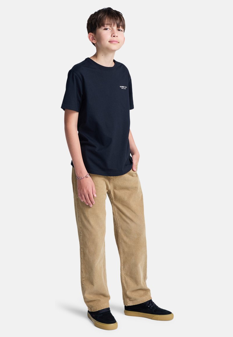 Garçon debout, mains dans les poches, portant un t-shirt noir, un pantalon beige et des baskets noires avec des semelles couleur gomme, sur fond blanc.