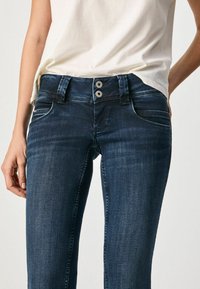 Jeans de mezclilla azul con cierre de doble botón, combinados con una camiseta blanca metida por dentro, con las manos descansando cerca de las caderas.