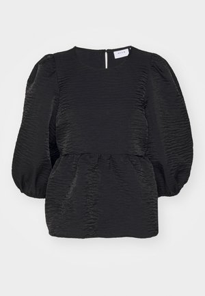 Femme portant un chemisier noir à manches bouffantes, un pantalon blanc fuselé et des mules à talons ouvertes noires, debout devant un fond gris uni.