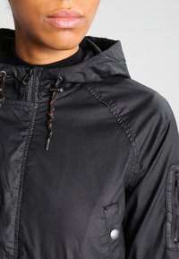 Veste légère noire avec fermeture à glissière sur le devant, capuche et cordons ajustables. Comprend une poche latérale zippée et un tissu texturé.