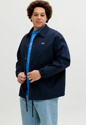Mand med krøllet hår iført en mørkeblå knap-op jakke over en blå skjorte og lyseblå jeans, står foran en helt hvid baggrund.