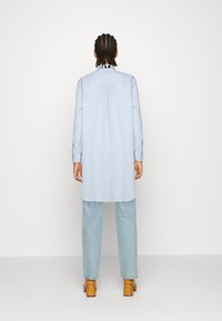 Person med flätat hår som bär en ljusblå lång tröja, blå jeans och bruna klackstövlar, stående med ansiktet riktat mot en vit vägg.