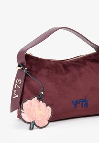 Bordeaux suède handtas met een zachte textuur, gouden rits en een geborduurd blauw logo. Voorzien van een roze bloemencharme die eraan bevestigd is.
