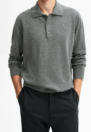Mann trägt ein graues langärmliges Strickpolohemd mit zwei Knöpfen und schwarze Hosen, Hände in den Taschen, vor einem weißen Hintergrund.