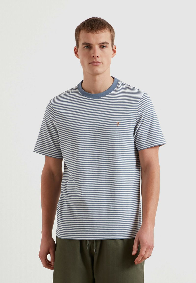 Jeune homme portant un t-shirt rayé bleu et blanc à manches courtes et un pantalon vert olive, debout devant un fond clair uni.