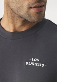 Maglietta in cotone grigio scuro con scollatura tonda, caratterizzata da un ricamo bianco e viola con la scritta "LOS BLANCOS" sul petto.