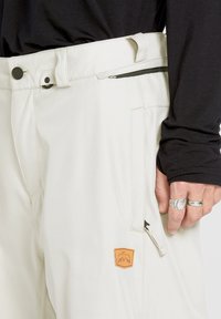 Pantalon beige clair à coupe décontractée, doté d'une poche zippée, d'une fermeture à bouton et d'un patch orange avec un logo sur le côté.