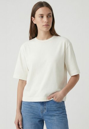 Młoda kobieta z długimi brązowymi włosami, ubrana w prostą białą koszulkę z krótkim rękawem i niebieskie dżinsy, stoi z jedną ręką w kieszeni.