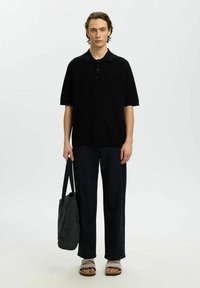 Jeune homme portant un polo noir à manches courtes, un pantalon foncé, des sandales grises, tenant un sac fourre-tout sombre sur un fond clair uni.